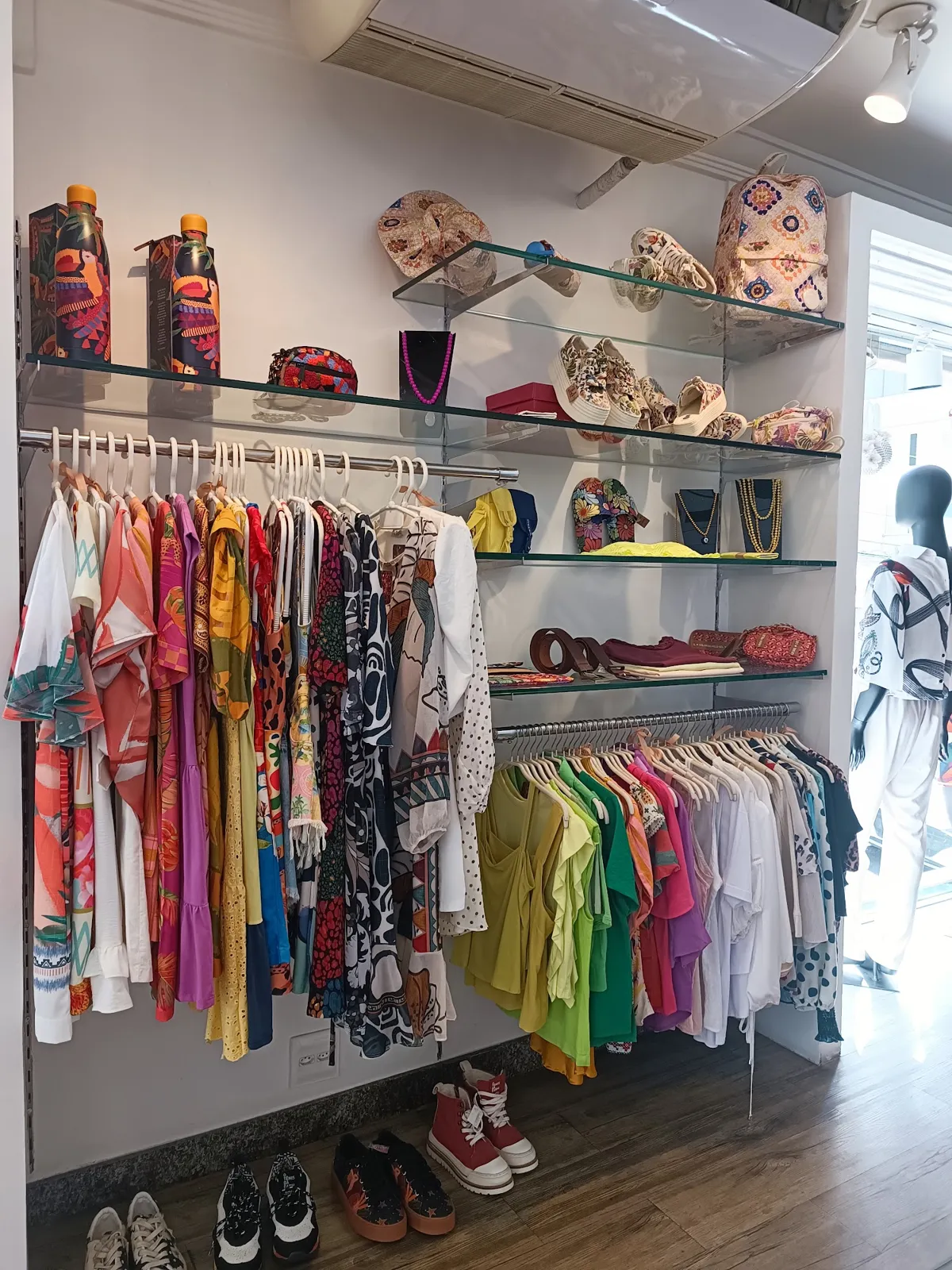 Blusas em cabides e prateleiras de vidro com bolsas e acessórios, dentro da Virtut